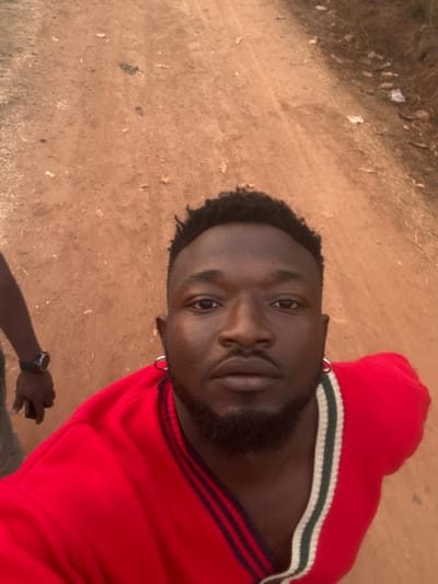 The image is a portrait of a young adult Black male, captured outdoors in Agbogugu, Nigeria. The man, seen from the chest up, has short, dark, curly hair and a neatly trimmed beard and mustache. He is looking directly at the camera with a calm, neutral expression. He wears a red sweater with white and dark blue stripes around the collar and cuffs, and small silver hoop earrings in both ears. A dark-colored wristwatch is partially visible on his left wrist towards the bottom of the frame.
The background consists of a reddish-brown, dusty ground, indicative of a dirt or sandy outdoor area, with some scattered dry vegetation and debris. The lighting suggests it is daytime, likely sunny, with natural light illuminating his face. There is no visible text in the image.