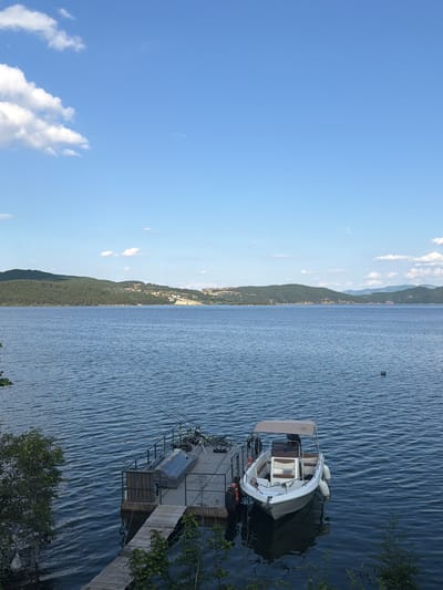 The image depicts a serene lake scene with a clear blue sky and scattered white clouds. In the foreground, a wooden dock extends into the calm, blue water. A small white motorboat with a tan canopy is moored to the dock, alongside other items such as life preservers and fishing rods. The water is rippled, suggesting a gentle breeze. In the background, a lush green, tree-covered hillside rises from the water's edge, dotted with houses and buildings, indicating a settlement. Further in the distance, more rolling hills are visible, creating a picturesque landscape. The overall impression is one of tranquility and natural beauty, likely a popular spot for recreation and relaxation in Zvanika, Bulgaria.