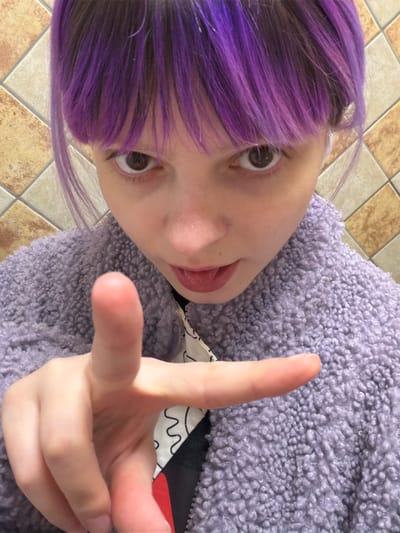 The image captures a close-up, high-angle perspective of an individual located in the city of Butsenino, Russia. The subject has fair skin, wide-set brown eyes, and bright purple hair with darker roots, cut with bangs. They are wearing a light purple, textured, sherpa-style jacket over an inner garment which is white with black abstract patterns and a black section, with a hint of red. The individual's mouth is slightly agape, revealing the tip of their tongue. Their right hand is raised towards the camera, with the index finger extended and pointing forward. The background consists of light beige or light brown square tiles with darker grout lines, arranged diagonally on a wall.