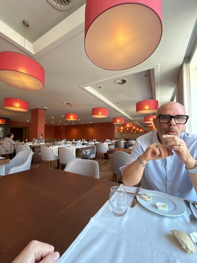 The image displays the interior of a restaurant, identified as being within Hotel Casino Chaves in Chaves, Portugal. In the foreground, a bald male with dark-rimmed glasses, wearing a light blue striped button-up shirt, is seated at a table on the right. His hands are clasped near his mouth, and he wears a gold-colored ring on his right ring finger, multiple bracelets on his left wrist, and a watch on his right wrist. On the white tablecloth in front of him are an empty stemless wine glass, silverware (fork, knife, spoon), a white plate containing a small piece of bread and a plastic container of butter, and a larger piece of bread. The photographer's left hand rests on the dark brown wooden table surface in the bottom left corner. The dining room is spacious, featuring numerous dark brown wooden tables, many set with white tablecloths, and light beige upholstered dining chairs. The white ceiling incorporates perforated acoustic panels and ventilation grilles, with multiple large, circular, red-orange fabric lampshades suspended, casting a warm light. The walls are painted an orange-tan color. Large windows are visible along the right side of the room, admitting natural light. In the mid-ground and background, other patrons are visible seated at tables, including a woman with curly hair and a patterned top on the far left, and a man seated alone further back.