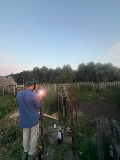 In the village of Kuseevo, Russia, a person is welding metal posts in an outdoor setting at dusk. The individual, wearing a blue shirt, beige pants, a dark baseball cap, and a welding helmet with yellow protective lenses, is standing on a small platform. Sparks are visibly emanating from the welding torch as it is used on a metal pole. The environment is rural, with a wooden fence, overgrown grass, and trees in the background. A wooden structure resembling a shed or outbuilding is visible to the left. The sky is a soft blue, transitioning to a lighter hue near the horizon. A welding machine and cables are on the ground nearby.