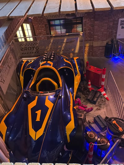 The image depicts a blue and yellow racing vehicle with the number '1' prominently displayed on its side. The vehicle appears to be a prop or a display, situated within an enclosed area with a metal fence. The environment suggests a workshop or a backstage area, with tools, cones, and other equipment scattered around. A red toolbox is visible near the vehicle. The background features brick walls and a window with horizontal blinds. In the upper portion of the image, wooden beams are visible, suggesting a ceiling structure. The overall impression is one of a themed exhibit or a prop display. The location in the city of Rust, Germany, is likely Europa-Park, given the nature of the exhibit.