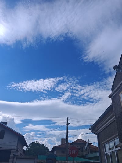 The scene is an upward-angled view captured in Tsarev Brod, Bulgaria, primarily depicting a dynamic sky and the upper sections of residential structures. The sky occupies approximately two-thirds of the image, showcasing deep blue areas interspersed with three distinct cloud layers: diffuse, high-altitude white clouds dominate the upper left and right, a clear band of blue sky stretches horizontally across the mid-upper section, and a more defined, broken band of white altocumulus clouds extends horizontally below the clear blue. A minuscule, dark, winged object, likely an aircraft or bird, is visible against the clouds in the upper right quadrant.
The lower third of the image frames portions of buildings and utility infrastructure. On the far left, a section of a light-colored house with a dark-tiled pitched roof and an upper-level window is partially visible. In the middle ground, a dark utility pole rises, supporting multiple black overhead power lines that crisscross the frame horizontally, extending between buildings. A red octagonal STOP sign with white lettering is prominently positioned in the lower-middle, mounted on a pole and partially obscuring the front of a background building. This background building features a reddish-brown tiled, pitched roof, a visible chimney, and a circular satellite dish affixed to its facade. On the far right, a dark grey, textured building facade is present, revealing a white-framed window on the lower right and a dark, wall-mounted outdoor lamp near its top edge. Dense green tree foliage is visible in the background, positioned between the structures. No people are visible within the frame.