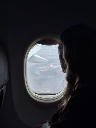 The image shows the interior of an aircraft with a view looking out an oval-shaped window, located over the city of Zuevo, Russia. A single individual is positioned in the right foreground, primarily in silhouette, with long, light-colored, wavy hair partially illuminated on their right side. The person is turned to the left, observing the external environment through the window. The aircraft cabin is dimly lit. Through the window, a white aircraft wing is visible, extending from the upper left into the frame. Below the wing, the ground consists of an industrial complex featuring multiple light-colored buildings, storage tanks, and associated infrastructure. Linear features suggestive of roads or power lines are visible across the landscape, which appears to be sparsely vegetated or barren under a hazy sky. The action depicted is an individual viewing the ground from an airborne aircraft.