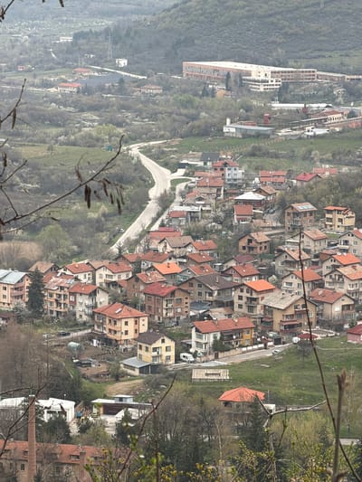 The image depicts a high-angle view of a residential area, characterized by densely packed houses with orange and brown tiled roofs, nestled amongst trees and greenery. A road winds through the landscape and the background shows hills and a mountainous region. The image appears to be taken in daylight, with an overexposed and slightly hazy atmosphere. Based on the description, the location is likely Bratsigovo, Bulgaria. The scene has a mix of residential buildings, foliage, and infrastructure, as one might observe from a vista point. There are no visible people or interactions in the captured perspective.