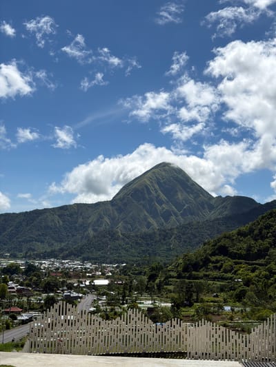 A photograph shows a verdant, mountainous landscape adjacent to a densely populated area under a sky with scattered clouds. The location is within the city of Sembalun Bumbung, Indonesia.
The mountain is covered in lush green vegetation, exhibiting visible terrain undulations. Below, a cluster of structures, likely buildings and residences, forms the populated area. Roads meander through the built environment.
The sky is bright blue, interspersed with white cumulus clouds. A man-made structure is visible at the right of the frame, appearing to be a wall or fence. No individuals are directly visible in the frame.