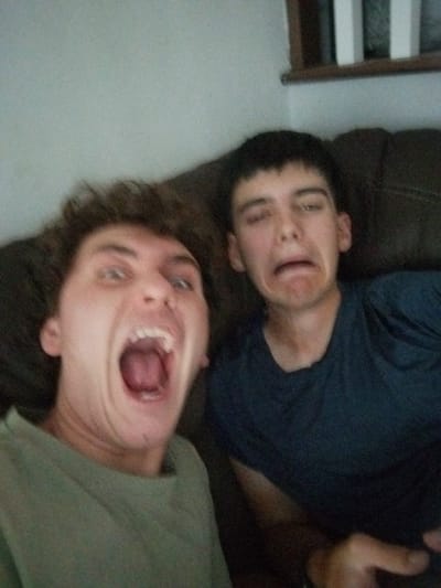 This indoor image shows two young adult males posing for a photograph, likely a selfie, within a residential setting. The individual on the left, positioned closer to the camera, has light brown, wavy hair and is wearing a light olive-green crew neck t-shirt. His mouth is wide open in an exaggerated expression, and his eyes are wide. The individual on the right, seated slightly behind and to the side, has short, dark hair and is wearing a dark blue crew neck t-shirt. His eyes are squinted, and his mouth is pulled downward in an exaggerated grimace. Both individuals are looking towards the camera. They appear to be seated on or against a dark brown upholstered couch or armchair, which occupies the mid-background. In the upper right background, a light-colored wall is visible, along with a partial view of what appears to be a white wooden balustrade or railing and a dark horizontal element, suggesting an interior space such as a living room. A hand is partially visible in the bottom right corner, possibly holding the device capturing the image. The precise location within Saint-Jean-sur-Richelieu, Canada, cannot be determined from the visual information provided in the image.