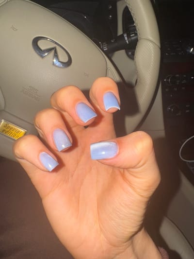 A person's hand with light blue, shimmery, square-shaped fingernails is held up in front of a car's steering wheel and dashboard. The steering wheel is a light beige color with stitching and the Infiniti logo in the center. The dashboard has various buttons and controls visible, along with a "SRS AIRBAG" label and a "Special Edition" sticker. The scene appears to be inside a car, possibly at night or in low light conditions, given the dark background and reflections. The hand is the primary focus, showcasing the well-maintained nails. There are no clear indicators of the weather or specific emotions being conveyed. The visible text includes "SRS AIRBAG" and "Special Edition". The location context provided is Новый Милет, Russia.