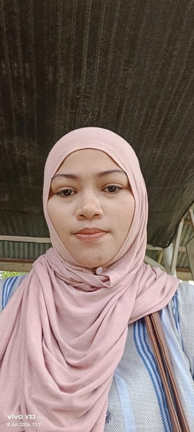 A medium shot selfie shows a woman wearing a pink hijab and a blue and white striped shirt. She is looking directly at the camera with a neutral expression. The background is blurred, but appears to be outdoors with some wooden structures and a dark, corrugated roof. The image has a timestamp in the lower left corner: "vivo V23 5G 8 Jun 2026, 13:11". The woman has light brown skin and dark brown eyes. She is wearing subtle makeup. The hijab is draped around her head and neck, obscuring most of her hair. She has a small brooch with a light-colored stone attached to the front of her hijab.