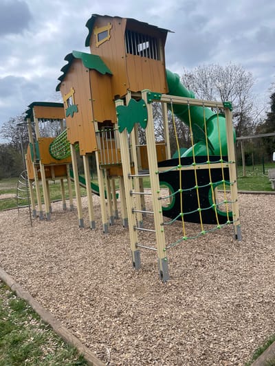 This image depicts a deserted outdoor playground in Holtzheim, France, under an overcast sky. The primary subject is a multi-component playground structure featuring a series of interconnected platforms and "treehouse"-like elements constructed from light brown, wood-patterned panels with dark green leaf-shaped accents on roofs and decorative elements. The structure is supported by numerous light-colored upright posts, some equipped with silver metal ladder rungs. Key features include a long, enclosed green tubular slide on the right, and a horizontal climbing tunnel with green and yellow netting. In the foreground, a vertical climbing wall is visible, consisting of dark panels with circular cutouts and a network of yellow and green climbing ropes. The ground directly beneath and surrounding the playground equipment is covered with light-colored wood chips, bordered by a circular wooden log in the immediate foreground, separating it from an area of green grass. In the background, deciduous trees with bare branches are visible, and a white park bench can be faintly seen on the right. No individuals are present in the scene.