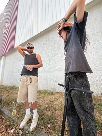 The image shows two individuals standing outdoors. The person on the left, wearing a black tank top, tan shorts, and a baseball cap, holds a clear bottle. The person on the right, in a dark hoodie and patterned pants, stands next to a scooter. They are positioned in front of a building with white and red vertical siding. The ground is covered in dry grass and fallen leaves. The scene appears to be in an urban or suburban setting. Based on the style of the building and the presence of urban elements, this location could plausibly be Caluire-et-Cuire, France, a commune on the outskirts of Lyon.