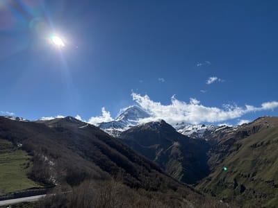 The image captures a daytime view of the mountainous landscape near Gergeti, Georgia. The sun shines brightly in the upper-left corner of the frame, creating a lens flare effect. In the center, a prominent snow-capped mountain peak rises above the surrounding ridges, partially obscured by a layer of white clouds. The lower slopes of the mountains are covered with dark, dense forests, while patches of snow are visible on the higher elevations. A road snakes along the base of the mountains in the lower-left corner of the image. The sky is clear blue with scattered white clouds.