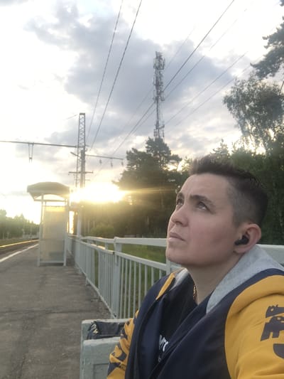 This media file is a portrait of a person at a train station in Tsibino, Russia. The subject, a person with short, dark hair and visible stubble, is wearing a dark blue and yellow jacket and an earbud. They are leaning on a railing, looking upwards and to the left. The setting is an outdoor train station with a concrete platform and a metal railing. In the background, there is a bright sun, suggesting it is either sunrise or sunset. The sky is partly cloudy. A telecommunications tower is visible in the background, along with power lines and trees. The overall mood is contemplative, with the subject seemingly lost in thought. No text is visible in the image.