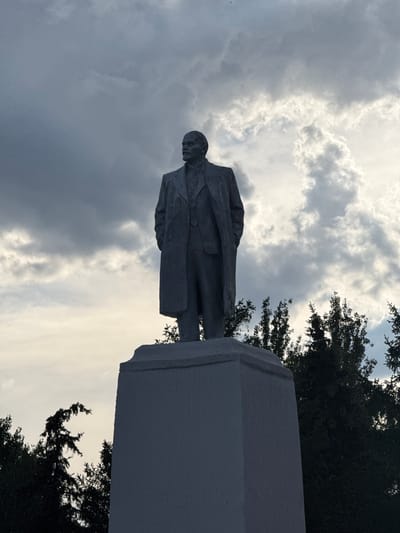 The media file depicts a statue of a man, likely a historical figure, standing on a pedestal. The statue is rendered in a grey, stone-like material and shows the man from the waist up, dressed in a suit and overcoat. He is facing left with his head turned slightly.
The scene is set outdoors against a dramatic sky filled with heavy, grey clouds, suggesting overcast or stormy weather. The lighting is dim, indicating it might be late afternoon or evening. To the right and upper left of the statue, dark green foliage from trees is visible, framing the sky and the monument.
The orientation of the image is such that the statue appears to be standing upright, with the pedestal at the bottom right of the frame and the statue extending upwards and to the left. The pedestal is a substantial, angular structure.
There is no visible text in the media file. The overall mood conveyed is somber and perhaps a bit imposing, due to the statue's presence and the heavy, cloud-filled sky. The location context provided, Masalovka, Russia, suggests a possible setting for such a monument. The image focuses on the statue and its immediate surroundings, highlighting the contrast between the man-made monument and the natural elements of the sky and trees.