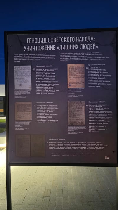 The image shows an outdoor information panel displaying information about the genocide of the Soviet people during World War II, specifically focusing on the extermination of "superfluous people" (those deemed undesirable by the Nazi regime). The location is likely a memorial or historical site. The panel contains text in Russian and reproductions of historical documents related to the persecution and extermination of disabled people, mentally ill individuals, and others deemed unfit by the Nazi regime in occupied territories of the Soviet Union, such as the Smolensk and Krasnodar regions. The documents include reports, testimonies, and official communications detailing atrocities committed by German forces. The panel is placed in an outdoor location with a paved area in the background.