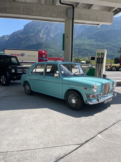 The image shows a light blue vintage car parked at a gas station with mountains in the background. The gas station has multiple pumps with branding partially visible. A red semi-truck and a black SUV are also present in the scene. The location appears to be a rest stop or service area along a highway. The vintage car has the license plate "EC-26-23." A concrete support structure, likely for a building above, is visible on the left. The lighting suggests it is a bright, sunny day. While Rans, Switzerland is a reasonable conjecture considering the truck with the company name "Spedler" and the Swiss license plate, the image does not explicitly show any signs indicating the city of Rans.