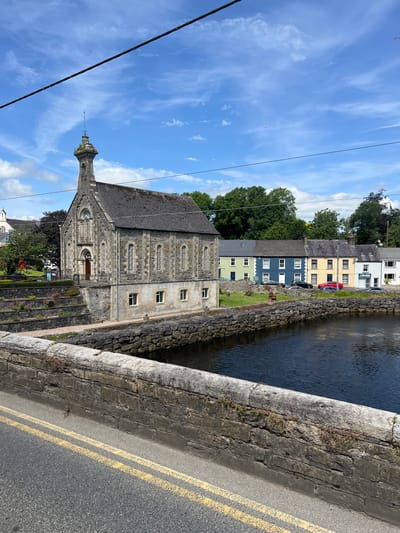 This is a vertical, eye-level, outdoor photograph taken on a sunny day with some clouds. The image captures a street scene in Donegal, Ireland, featuring a stone building, a body of water, and a road with double yellow lines.

The main subject is a long, grey stone building with a steep, dark roof. It has several arched windows with intricate stone detailing and smaller rectangular windows. A spire-like structure with decorative metalwork rises from the roofline near the front of the building. The building appears to be an older structure, possibly a historical or religious building.

To the right of the stone building is a body of dark, calm water, possibly a river or canal. Its edge is lined with rough stone walling. Beyond the water, a row of colorful terraced houses is visible. These houses are painted in shades of yellow, blue, and green, with white window frames. Some cars are parked in front of them.

In the foreground, a stone bridge or embankment runs diagonally across the image, separating the road from the water and the building. The road itself has double yellow lines painted on it, indicating a no-overtaking zone.

The sky is a bright blue with scattered white clouds, some wispy and others more defined. Two birds are visible flying in the sky. The overall impression is of a picturesque and tranquil village or town scene. The time of day appears to be daytime, likely late morning or early afternoon, given the bright sunlight casting shadows and illuminating the buildings. The weather is fair and pleasant.