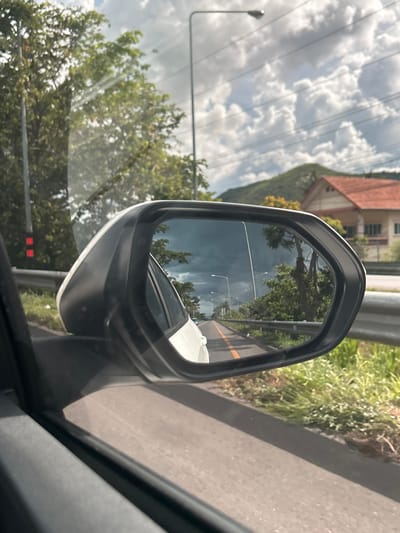 This media file shows a view from inside a car, looking towards the side mirror. The side mirror reflects a scene of a road with lush green trees and a cloudy sky. The road appears to be a two-lane road with a yellow line separating the lanes. Streetlights are visible along the road in the reflection. In the background of the reflection, there's a white car and some trees.

Outside the mirror, the viewer sees the car's interior and the surrounding environment. A grey metallic guardrail runs along the side of the road. Green vegetation, including trees and bushes, is abundant. The sky is filled with dramatic, puffy white clouds against a light blue backdrop. A building with an orange tiled roof is visible in the upper left portion of the frame, suggesting a semi-rural or suburban setting. A streetlight is also visible to the left of the mirror.

The overall scene suggests a drive in Ao Noi, Thailand, possibly during the daytime given the bright sky. The clouds indicate potentially changeable weather. The image captures the typical scenery of a roadside environment in Thailand, with a focus on the reflection in the car's side mirror. There is no visible text in the image.