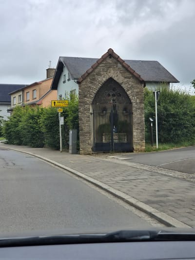 The image depicts a street scene in Weiler-la-Tour, Luxembourg. In the foreground, a grey road curves to the right, with a paved sidewalk bordering it. Beyond the sidewalk, a row of dense green hedges lines the street. To the right of the hedges, a stone structure with an arched entrance and a metal gate stands prominently. The gate is ornate, with intricate ironwork, and appears to enclose a small garden area with plants.
Behind the hedges and the stone structure, several houses are visible. These include a two-story house with a light blue facade and a grey roof, a peach-colored house with a grey roof, and another house with a grey roof and solar panels. A prominent yellow street sign is mounted on a pole, indicating "HASSEL" with an arrow pointing to the right, and "WEILER-LA-TOUR" below it, along with the number "132". Another smaller sign is also visible on the same pole. The sky is overcast with a grey, cloudy atmosphere. The overall scene suggests a residential or semi-rural setting.