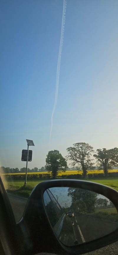 The image shows a view from inside a vehicle, likely a car, looking out towards a roadside scene. The location is Broadland, United Kingdom. The sky is blue with a contrail visible. A field of yellow flowers runs alongside the road, with several trees dotting the landscape. A roadside marker with a solar panel is visible. The vehicle's side mirror reflects the road behind, showing a car and other vehicles.