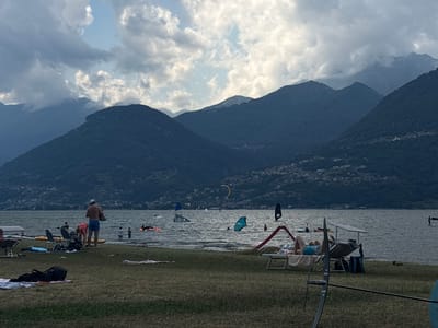 The image depicts a lakeside scene in Colico, Italy, with a focus on the water and the surrounding mountains. The water is choppy, and several individuals are engaged in water sports, including windsurfing and kitesurfing, with visible sails and boards on the lake. In the foreground, there is a grassy area with several people relaxing on the shore. Some are sitting in lounge chairs, while others are lying on towels. There are also various beach accessories present, such as bags and a beach umbrella. The background is dominated by a range of lush green mountains that rise steeply from the lake, with a small town or village visible on the mountainside. The sky is partly cloudy, with patches of blue sky interspersed with dramatic grey clouds. The overall impression is one of a recreational day at the lake, with people enjoying the water and the natural beauty of the landscape.