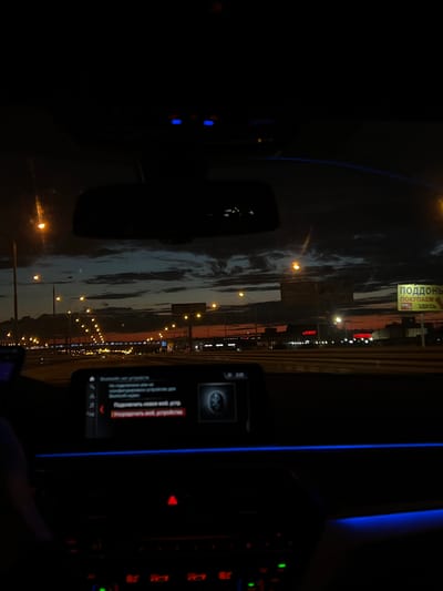 The image shows the interior of a car at night, with the driver's perspective looking out onto a highway. The sky is dark with dramatic clouds, illuminated by the orange glow of streetlights and the setting sun on the horizon. There are several billboards visible along the highway, one of which has yellow signage with Russian text. The car's dashboard is visible, with a screen displaying what appears to be a car's infotainment system, showing Russian text and a Bluetooth icon. The interior of the car is lit by subtle blue ambient lighting. The scene appears to be on a highway outside of a city, potentially near an industrial or commercial area, given the billboards and buildings visible in the distance. No people are clearly visible in the frame, though a hand can be seen in the lower left corner.