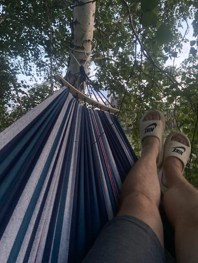 The image depicts a person lying in a hammock suspended between two trees. The hammock is made of striped fabric in shades of blue, white, and grey, with a red stripe visible. The person's legs are visible, wearing grey shorts and white slide sandals with the Nike logo. They are positioned with their feet up, resting on the hammock. Surrounding the hammock is lush green foliage from the trees, and the sky is visible through the leaves. The scene suggests an outdoor setting, likely a park or backyard, in the city of Сухая Балка, Russia. The lighting indicates it is daytime, possibly late afternoon or early evening, with soft light filtering through the trees.