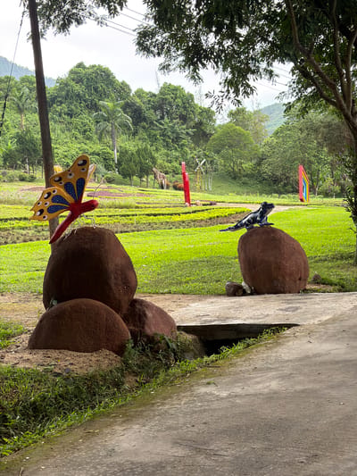 The image depicts an outdoor scene in Khánh Vĩnh, Vietnam. In the foreground, a pathway made of concrete leads into the scene. To the left, a cluster of large, brown, rounded rocks is visible, with a colorful butterfly sculpture perched on the top rock. The butterfly has orange wings with blue and black markings. To the right of the pathway, another large brown rock supports a black and white striped lizard sculpture.
The middle ground of the image features a meticulously maintained green lawn with rows of plants. In the background, lush green trees and hills dominate the landscape, suggesting a rural or park-like setting. Several colorful, abstract sculptures are scattered throughout the lawn, including a red pole, a yellow windmill-like structure, and a red and blue geometric sculpture. A distant giraffe sculpture is also visible. The sky is overcast, contributing to the diffused lighting.