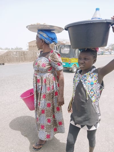 Two individuals, an adult female and a younger female, stand on a paved surface under a bright sky in Kadoma, Zimbabwe. The adult female, positioned to the left, wears a long, patterned dress and a blue headwrap. She balances a silver-colored, shallow metal tray containing light-colored items on her head and carries a pink plastic bucket in her left hand. She is looking to her left. The younger female, to the right, wears a black and multi-patterned sleeveless top with black leggings. She balances a large black plastic basin, containing a blue plastic bottle, on her head, supporting it with her raised left arm. She is looking forward, towards the viewer. In the mid-ground, a yellow and light-blue three-wheeled auto-rickshaw travels leftward on the road. Faint structures and a dusty environment are visible in the background under strong sunlight, casting shadows on the ground.