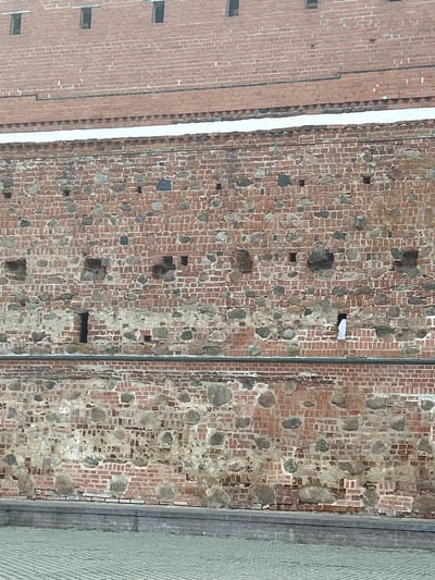 The image shows a weathered brick wall with embedded stones and small openings. The wall is constructed in sections, with the upper part featuring uniformly laid red bricks and narrow, dark openings. Below this, a wider section of the wall is visible, characterized by a more irregular pattern of red bricks interspersed with larger, rounded stones of varying shades of gray and brown. Some areas of the brickwork in this section appear damaged or worn, revealing lighter mortar underneath.
Further down, there is another horizontal band of brick and stone construction, appearing similar in style to the section above it. The bottom of the image shows a paved area and a stone ledge or base for the wall. The overall impression is of an old, robust structure, possibly a fortification or ancient building, located in an outdoor setting. The lighting suggests it is daytime, with no visible people or specific landmarks that would pinpoint the exact location within Жухавіцкі сельскі Савет, Belarus.