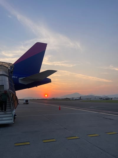 The image captures a scene at Orio al Serio International Airport in Italy. A Wizz Air Airbus A321 aircraft, distinguished by its magenta and dark blue tail, is docked with an airport jet bridge. Passengers are visible entering or exiting the aircraft via the jet bridge. The setting sun casts a warm glow across the sky, with visible linear cloud formations. An orange traffic cone is placed along the edge of the tarmac. Other aircraft, one of which is identifiable as a Ryanair aircraft, are parked further down the tarmac. The airport infrastructure, including painted yellow markings on the tarmac, are visible. Mountains are present in the distance, adding to the background. The overall scene presents an evening flight or arrival at the airport.