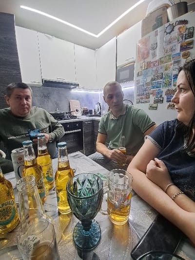 A group of people are gathered around a table in a kitchen. There are bottles of beer and filled glasses on the table. One person is pouring beer into a glass. The kitchen has white cabinets and a modern design. There are magnets and other items attached to the refrigerator.