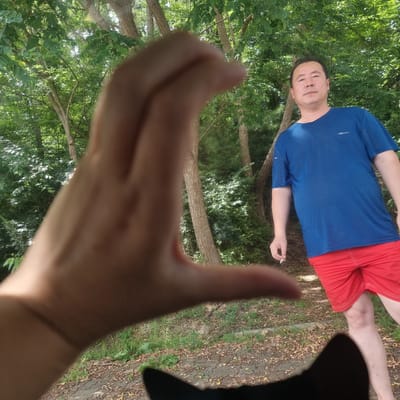 A man is pictured in a wooded area. He is wearing a blue striped t-shirt and red shorts. The man is holding a cigarette in his right hand. The background consists of trees and foliage. A hand is in the foreground, obscuring part of the image. The scene appears to be outdoors, likely in a park or hiking trail, in Gangneung-si, South Korea.