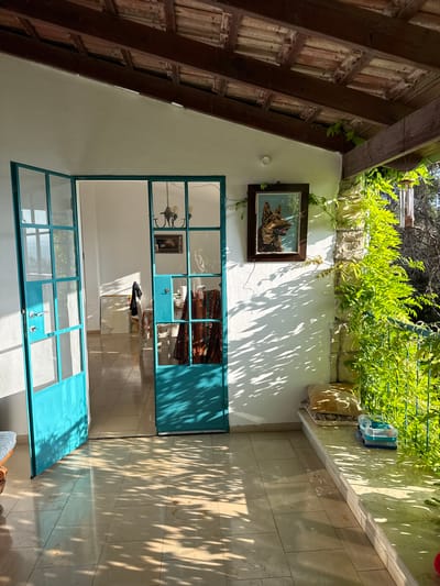 The image depicts a sun-drenched patio area with a covered roof in Kfar Vradim, Israel. Two teal-colored glass doors are open, revealing an interior space with tiled floors and white walls. Sunlight casts dappled shadows from foliage onto the patio floor and walls.
On the patio, a stone ledge runs along the right side, adorned with two cushions and a blue and clear plastic container. Lush green vines climb the stone pillar and hang over the ledge, suggesting a garden setting. A wind chime is visible hanging from the wooden roof structure.
The interior visible through the doors features tiled floors, white walls, and a chandelier. A framed picture of a dog hangs on the exterior wall, to the left of the right door. The overall impression is one of a relaxed, outdoor living space bathed in natural light.