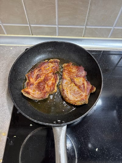The scene is located in a kitchen within the city of Mikrevo, Bulgaria. A black non-stick frying pan occupies the central foreground, resting on a black ceramic stovetop. Inside the pan, two irregular pieces of browned meat, consistent with pork or beef steaks, are observed cooking in a quantity of translucent yellow oil. Visible signs of cooking include browning on the meat surfaces and small bubbles in the oil. The stovetop shows minor residue. In the background, a light grey countertop extends horizontally, surmounted by a tiled backsplash consisting of light beige square tiles with dark grout. A thin, horizontal light grey strip is positioned between the countertop and the tiled surface.