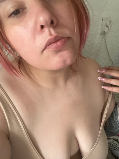 The image shows a close-up, chest-up view of a person with light skin and pink-tinged hair. The person is wearing a light-colored, possibly beige, tank top. The focus is on their upper chest, shoulders, and the lower part of their face, including their lips and nose. Their right hand, with French manicured nails, is resting on their right shoulder. In the background, a white electrical outlet with a plug is visible on a textured wall. The lighting appears to be artificial and somewhat dim. There is no clear indication of the location being in Коробицыно, Russia, or any specific city.