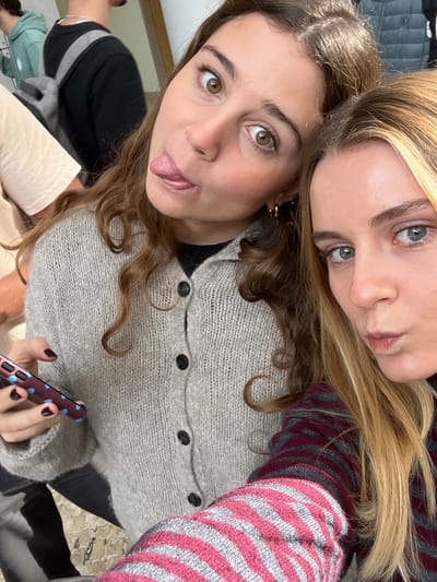The image depicts two women in the foreground, with several individuals partially visible in the blurred background, within an indoor setting in Porto Salvo, Portugal. The woman on the right has light brown, wavy hair and brown eyes; she extends her tongue and widens her eyes. She wears a light grey knitted cardigan with five visible dark buttons over a dark garment, along with gold hoop earrings. Her left hand, with dark red painted fingernails, holds a smartphone featuring a pink case with blue polka dots. To her left, a woman with long blonde hair and light-colored eyes is partially visible, pouting her lips. She wears a horizontally striped long-sleeve shirt in dark red and grey tones. In the background, to the far left, a person wearing a light green hooded sweatshirt is partially visible from the back. Another individual, wearing a black long-sleeve shirt and a grey backpack, is also partially visible behind the brown-haired woman. The visible environment includes a light-colored wall and a light stone or tiled floor in the lower left.