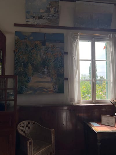 This image depicts an interior room, specifically the studio or dining room of Claude Monet's house in Giverny, France.

The scene focuses on three distinct oil paintings displayed on a light-colored wall above dark wooden wainscoting. The most prominent painting, centrally located and approximately square, illustrates a sunlit garden path lined with vibrant sunflowers, leading up to a house. A person in a light-colored hat walks along the path in the foreground, with another figure further up. Above this, on a dark wooden shelf, two smaller paintings are visible: one on the left shows a rocky coastline with boats on a beach under a cloudy sky, and another on the right is a landscape with a muted sky and distant structures.

To the right of the large painting, a multi-pane casement window, framed in white, offers a view of a lush garden outside. Sheer white curtains are tied back on either side of the window. The exterior scene reveals dense green foliage, various flowering plants (including red and purple blossoms), and a tall, slender tree. Bright daylight illuminates the outdoor scene, contrasting with the slightly dimmer interior. A red plant or flower is visible near an open pane of the window on the right.

The lower half of the room features dark, polished wooden paneling. In the bottom left quadrant, a natural wicker armchair with armrests is positioned. To the right, beneath the window, a small, dark wooden table with an inset reddish-brown top stands, bearing a small, framed informational plaque. On the far left, a dark wooden cabinet or doorway with glass panes is partially visible, containing dimly lit ceramic vessels.