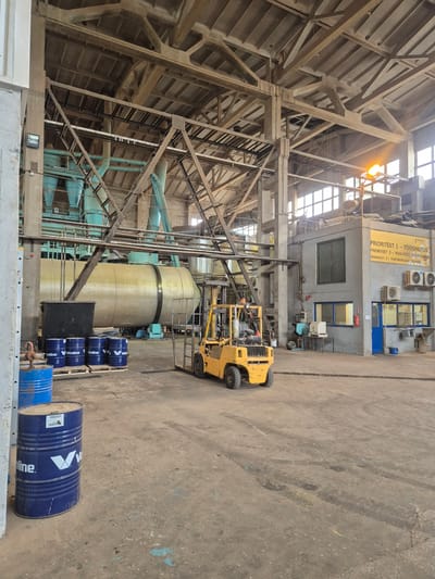 This image depicts the interior of an industrial building, likely a factory or warehouse, in Ebavere küla, Estonia. The scene is dominated by large, industrial machinery, including towering cylindrical structures and extensive piping systems, rendered in muted tones of beige and teal. Wooden beams and metal scaffolding crisscross the ceiling and walls, suggesting a substantial construction.
In the foreground, several blue metal drums with white lettering, some stacked on wooden pallets, are visible. The visible text on these drums includes "Valvoline," indicating they likely contain industrial lubricants or chemicals. A yellow forklift truck is positioned near the machinery, suggesting ongoing or recent logistical activity within the space.
The lighting suggests it is daytime, with light filtering in from a large, high ceiling composed of segmented windows. The overall impression is one of an active industrial environment, possibly engaged in manufacturing or processing. A yellow sign with black text is mounted on a concrete wall in the background, bearing the heading "PRIORITEET" followed by numbered priorities and descriptions in Estonian: "TÖÖOHUTUS" (Work safety), "KVALITEET, EFFEKTIIVSUS" (Quality, Efficiency), and "TOOTMISPLAANI TÄITMINE" (Fulfillment of the production plan). This sign provides insight into the operational focus of the facility. The concrete floor is marked with tire tracks and some staining, indicative of regular use.