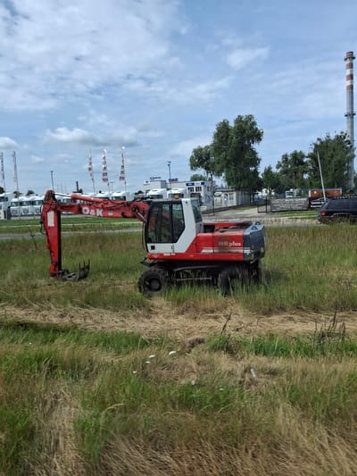 The image depicts an excavator in a grassy field. The excavator is red and white and has the brand "O&K" visible on its arm. In the background, there are buildings with flags, trucks, and a tall industrial chimney. A black SUV is parked near a building with signage that reads "AUTO SERWIS" and "WAGRA BL". The scene is set in Cierpice, Poland, under a cloudy sky. There are no people visible in the image.