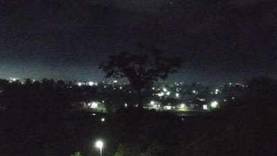 The image depicts a nighttime scene of a residential area in Blangtingkeum, Indonesia. The foreground is dominated by dark silhouettes of trees and foliage, with a prominent, large tree with a broad canopy standing in the mid-ground. Beyond the trees, a scattering of lights indicates houses and possibly streetlights, suggesting a village or suburban setting. The lights are a mix of warm yellow and cooler white hues, creating a soft glow against the dark landscape. The sky is dark, characteristic of nighttime, with a faint haze or mist visible in the distance, possibly illuminated by the lights of the settlement. There are no visible people, objects, or text in the image. The overall mood is serene and quiet, typical of a rural or suburban environment at night. The location cues, such as the arrangement of houses and the presence of lush vegetation, are consistent with a tropical or subtropical setting.