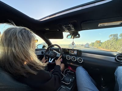 The image shows the interior of a Mercedes-Benz car driving on a highway. The car's sunroof is open, revealing a sunny, clear sky. A person with long, gray hair is behind the steering wheel, with both hands on it. The car's dashboard and console are visible, including the infotainment screen and air vents. Another person's legs, clad in blue jeans, are visible in the passenger seat. Other cars and greenery are visible through the windshield, suggesting the vehicle is on a highway. The image was likely taken in Huntington Beach, United States, according to the prompt instructions.