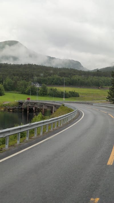 The image depicts a view of a road curving to the right, leading towards a bridge. The road is asphalt with a yellow line marking. A silver guardrail runs along the left side of the road. The bridge is concrete with visible supports and arches, spanning over a body of water. On the far side of the bridge, there is a red vehicle. The background features a lush green hillside with scattered trees and a few buildings, including a larger house with multiple windows. Further in the distance, mountains are visible, shrouded in mist. The sky is overcast with clouds. The scene is located in the municipality of Engan, Norway.