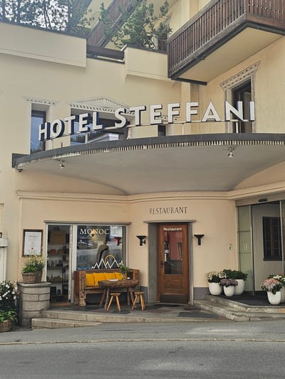 This is a photograph of the exterior of a hotel and restaurant in St. Moritz, Switzerland. The building is light-colored with dark trim and features a prominent sign that reads "HOTEL ST. EFFA NI". The hotel has a traditional facade with awnings and balconies.
In front of the building, there is a street with a sidewalk. The sidewalk features several decorative elements, including planters with flowers, a rustic wooden bench with yellow cushions, and a large barrel planter. A wooden door with a glass panel leads into the establishment, and next to it, a window displays the word "MONOCLE" and a menu. Above the door, the word "RESTAURANT" is visible.
To the left of the hotel entrance, a green tree with dense foliage is growing against the building. The time of day appears to be daytime, with overcast or diffused lighting, suggesting it might be a cloudy day. The overall impression is one of a well-maintained establishment in a European town.
Visible text includes "HOTEL ST. EFFA NI", "RESTAURANT", "MONOCLE", and "allegra".