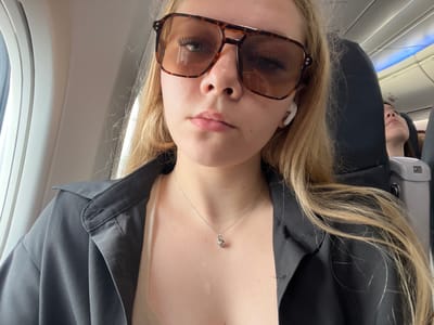 A young blonde woman, wearing large brown sunglasses, a light-colored camisole, and an unbuttoned black shirt, is seated on an airplane. She has her hair down and is wearing white wireless earbuds. A silver necklace with a small pendant is visible on her chest. The background shows the interior of an airplane cabin with a window to her left and another passenger in the seat behind her. The scene does not depict the city of Сокол, Russia, but rather the inside of an airplane.