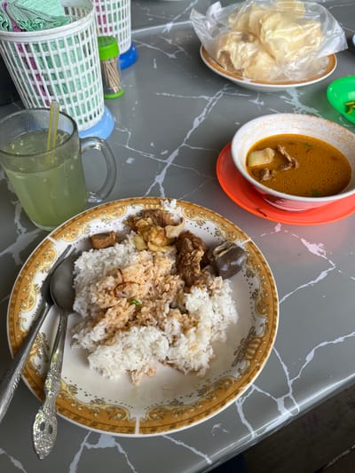A top-down view captures a meal served on a grey table with white marble-like veins, likely in Aluegunto, Indonesia. The main dish is presented in a decorative white and gold-patterned bowl, containing white rice mixed with a brownish-orange sauce or seasoning, along with several pieces of meat (possibly beef or goat) and a dark piece of offal. Two silver spoons rest in the bowl.
To the left of the main dish, a clear glass contains a greenish-yellow beverage with a straw, showing condensation on its surface. Above this glass, two white, lattice-patterned plastic holders are visible, one containing green folded tissues or napkins, and another with a small container of toothpicks. A small, bright green dish holds several green chili peppers.
Further up and to the left, a white bowl containing light-colored crackers, partially enclosed in a clear plastic bag, is visible. Adjacent to this, an off-white bowl with a similar brownish-orange soup and pieces of meat and a light-colored vegetable (possibly chayote or potato) rests on a bright orange saucer. The overall setting suggests a casual dining experience. There is no visible text in the image.