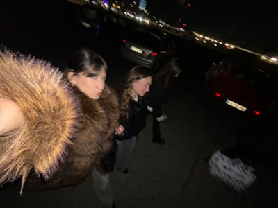 The image depicts an outdoor night scene in Lampertheim, France, captured with a significant clockwise rotation. The lighting is dim, relying on ambient sources and artificial lights.

In the foreground, a person is prominently featured, their face turned towards the viewer. They are wearing a substantial dark brown, possibly faux fur, coat with a prominent fluffy collar. Their dark hair is visible. To their left and slightly behind, another individual is partially visible, also looking towards the camera, with lighter hair and dark clothing. A third person in dark attire is faintly discernible further back in the scene.

The setting appears to be a street or parking area. A red car with its taillights illuminated is visible in the mid-ground to the left. A French license plate, displaying "KO 852-B", is visible on the rear of this vehicle. Another dark vehicle is present further in the background. The far background features numerous blurred lights, indicating distant traffic or urban illumination. A white and blue patterned object or fabric lies on the ground in the upper left portion of the image.

The scene suggests a casual gathering or encounter among friends outdoors at night. The orientation of the image and the direct gazes imply a candid photograph or a selfie. The fur coat suggests potentially cool or cold weather conditions.