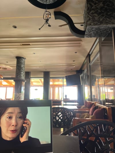 The image shows a laptop screen displaying a close-up of an East Asian woman speaking on a mobile phone. The laptop is placed on a table in what appears to be a restaurant or cafe with a rustic and somewhat formal interior design. The background is blurred, revealing seating areas with wooden furniture and plush cushions, large windows with orange drapery, and decorative pillars. The lighting suggests it is daytime, with sunlight streaming through the windows. The location is identified as El Hajeb, Morocco.