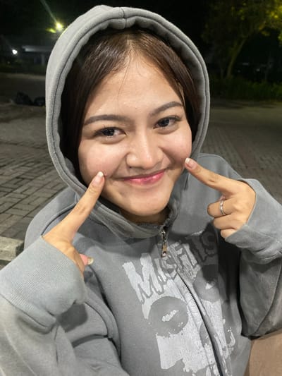 A young woman with brown hair, wearing a grey hooded sweatshirt, is captured in a close-up, outdoor shot. She is smiling and pointing at her cheeks with both index fingers. The background appears to be a paved area with some greenery and artificial lighting, suggesting an urban park or public space. The time of day appears to be evening or night due to the lighting conditions. The overall scene is casual and candid. The location is identified as Mekar Bakti, Indonesia.