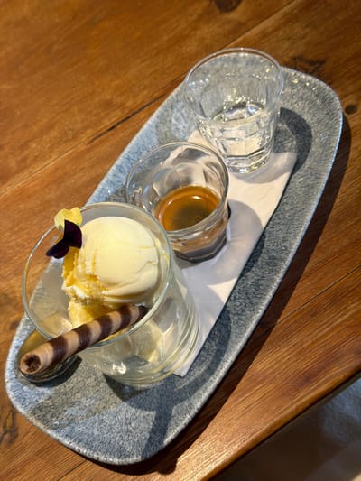An overhead shot captures a dessert and coffee arrangement presented on a wooden table. The items are served on a grey, textured, and irregularly shaped serving tray. On the tray, there are three distinct glasses: a large, clear, faceted glass containing water, a smaller glass holding a shot of dark brown espresso with a light brown crema, and a wider glass filled with a scoop of pale yellow vanilla ice cream. The ice cream is garnished with a striped chocolate wafer roll and a small, delicate flower with purple and yellow petals. A white napkin is positioned beneath the water and espresso glasses.
The underlying surface is a warm-toned wooden table, exhibiting visible grain. Bright, directional lighting from the top right casts distinct shadows from the glasses and tray onto the table and napkin, indicating daytime. The setup suggests a dessert or coffee service, possibly an affogato, being enjoyed in a cafe or restaurant setting in Theewaterskloof Nu, South Africa. No people are visible, and there is no discernible text in the image.